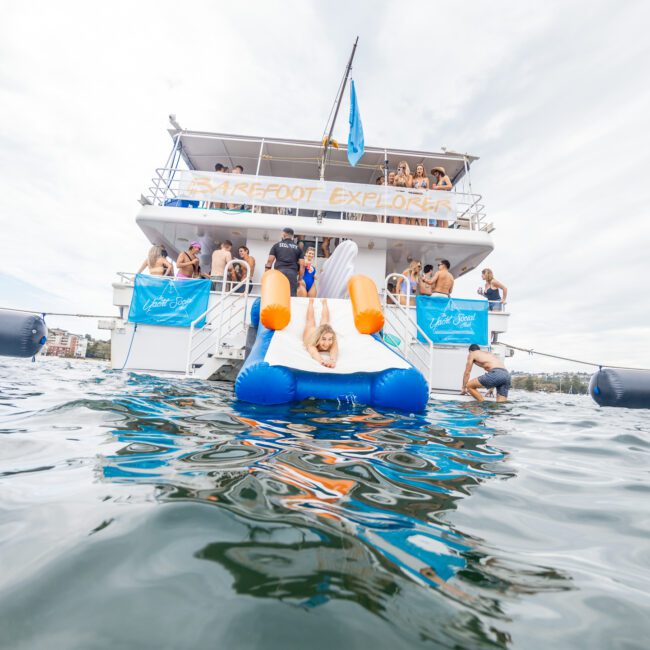 A person lounges on a large inflatable swan floating in the water near the rear of a double-deck boat named "Barefoot Explorer." The boat, brimming with enthusiastic people, showcases a lively scene. The background reveals a stunning coastline dotted with other boats.