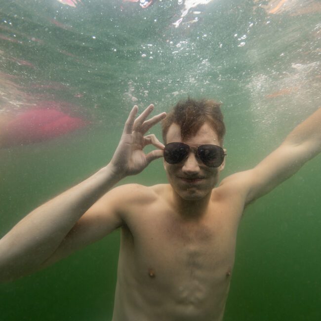 A shirtless individual underwater making an okay hand gesture with one hand and wearing dark sunglasses, surrounded by a greenish hue. Part of another person's body is visible on the left side of the frame. The text “The Yacht Social Club” is in the bottom right corner, evoking an exclusive vibe.