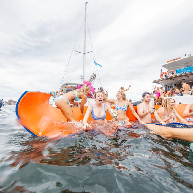 A group of friends is having fun on an orange floating mat in the water near anchored yachts. The sky is overcast, and everyone is enjoying themselves, smiling and cheering. Some are in the water while others relax on the mat, creating a festive atmosphere.