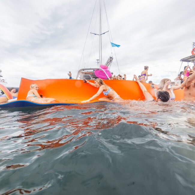 A group enjoying a lively party on the water, with some on boats and others using colorful floats. One person relaxes on a large orange float while others are nearby in the water. Several yachts add to the picturesque scene in the background.