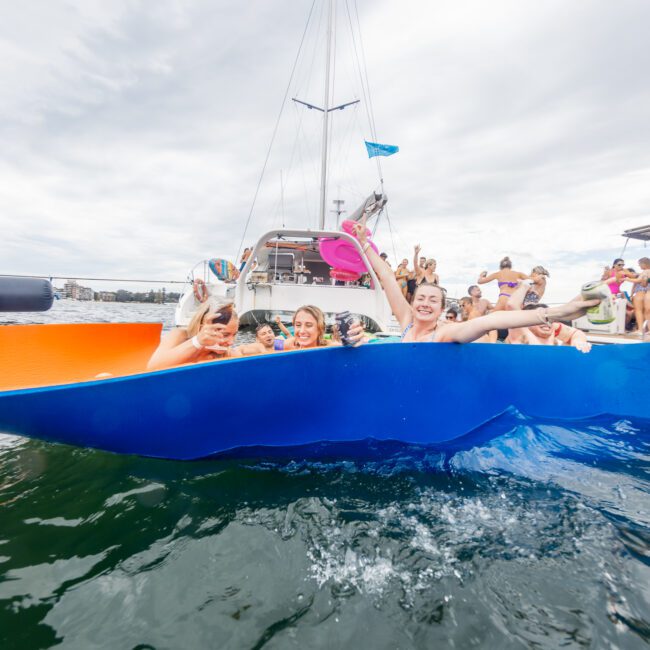 A group of people smile and wave from the edge of a vibrant catamaran in a body of water. Other boats with more people are seen in the background on an overcast day. Text in the bottom right corner reads "Yacht Social Club.