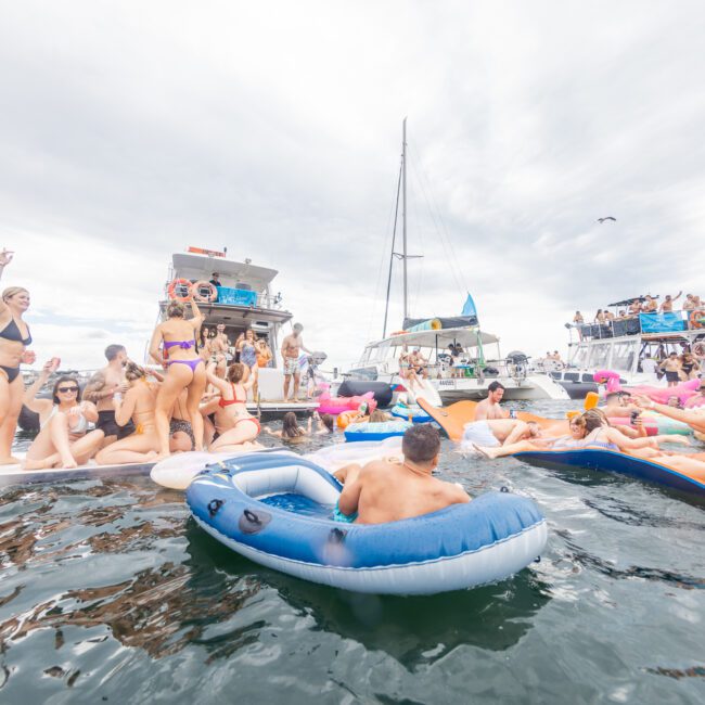 A lively group of people are enjoying a yacht party on the water. Some are dancing on the yacht while others relax on floating inflatable rafts and pool lounges. The sky is cloudy, and the water is calm, creating a perfect backdrop for the festive, social atmosphere.