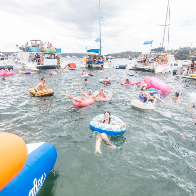 People enjoying a lively yacht party on the water, floating on inflatable tubes and rafts near anchored boats. The sky is cloudy, and the atmosphere is relaxed and festive. The water is calm, and some people are waving at the camera. Logo in bottom right corner reads "Yacht Social Club.