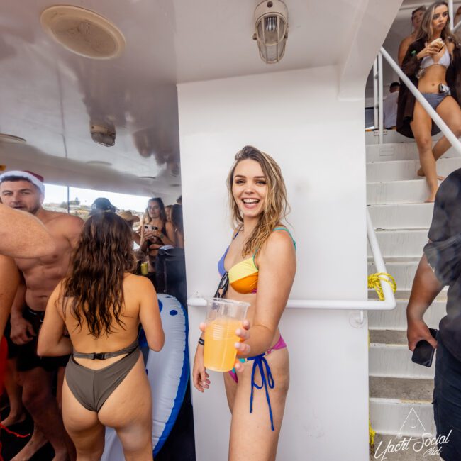 A smiling woman in a vibrant multicolored bikini holds a drink and stands near a stairway on a yacht during a lively party. Other partygoers in swimwear mingle in the background. The festive atmosphere buzzes as people relish the event.