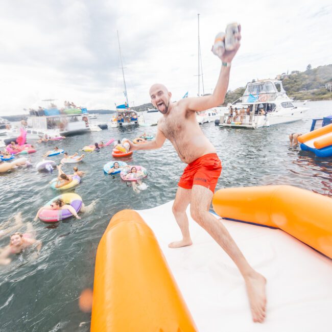 A man in red swim trunks stands on an inflatable platform, holding a case of drinks and grinning widely. Behind him, people float on colorful inflatables in the water, with boats anchored and a hill glowing under the sun in the background. The scene is lively and joyful.