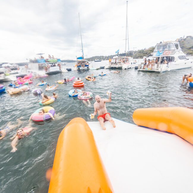 A lively scene of a group enjoying a day out on the water. Some are floating on various inflatable devices, including a swan and flamingo, while others swim. Boats are anchored nearby, and the sky is partly cloudy. One person slides down an inflatable slide into the water.