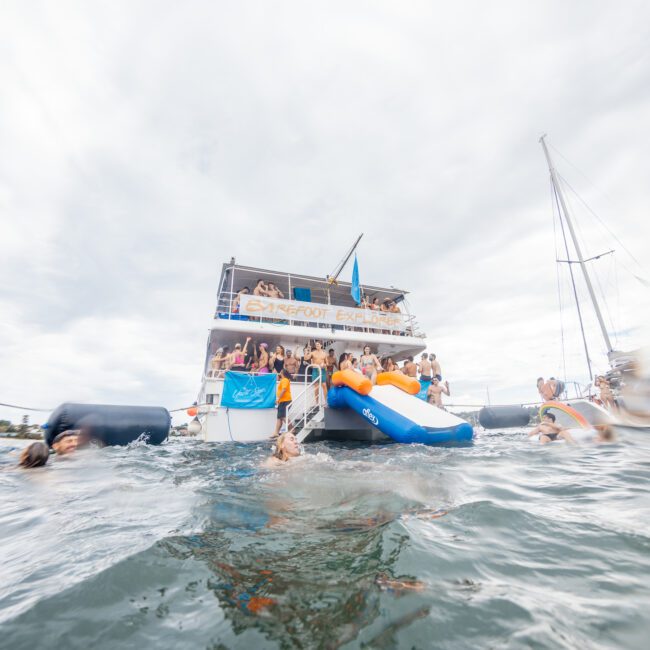 A lively scene of people enjoying a party on a large yacht with an inflatable slide leading into the water. Several individuals are swimming and interacting near the yacht, with another vessel visible nearby. The overcast sky creates a muted yet cozy backdrop as everyone revels in their maritime adventure.