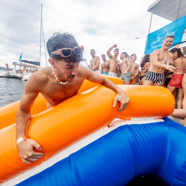 A man in swim trunks climbs out of the water onto an inflatable orange raft near a party boat. He's surrounded by people wearing swimsuits, some cheering and enjoying the festivities, with yachts and a sunny sky in the background. There is "The Yacht Social Club" logo in the corner.