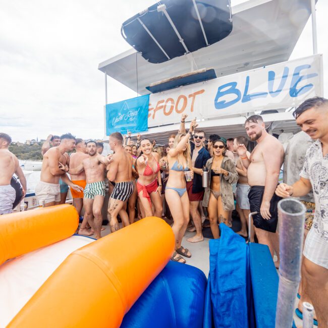 A large group of people in swimsuits are partying on a boat named "Foot Blue." They are gathered around smiling, laughing, and holding drinks. The scene is lively and festive with inflatable items dotting the deck and a stunning sunset in the background.