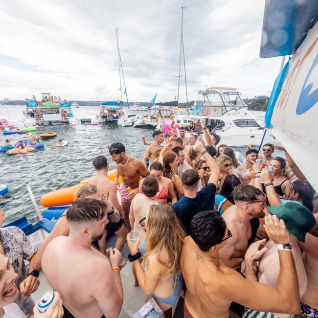 A lively group of people socializing on a boat during a yacht party. Many attendees, dressed in swimwear, are gathered closely together, enjoying drinks and music. Other boats and floating inflatable structures are visible in the background on the shimmering water.