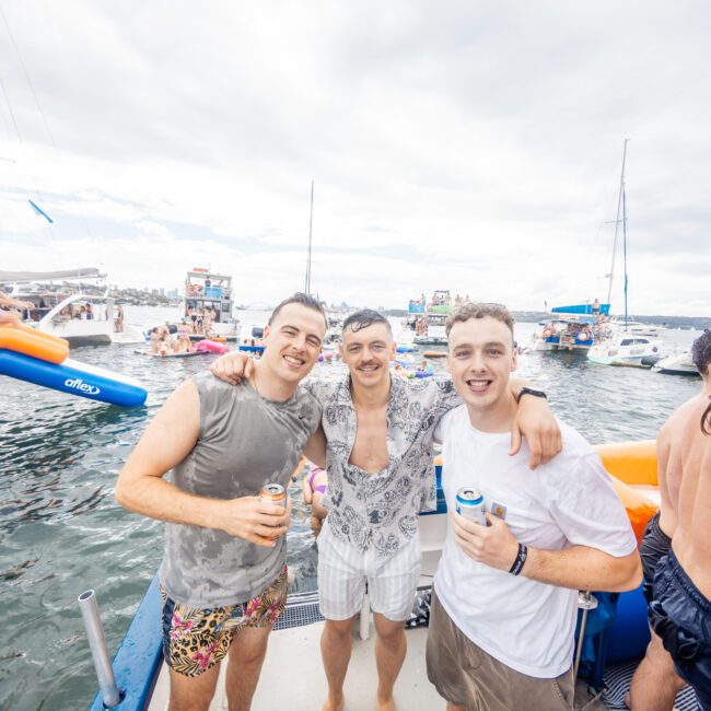 Three men smiling and posing on a boat in a lively water setting, holding canned beverages and wearing casual summer clothes. Surrounding them are other boats and people enjoying the water under a partly cloudy sky, creating a festive atmosphere.