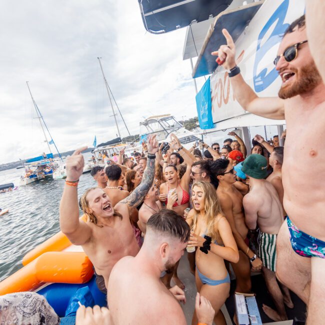 A group of people in swimwear having a lively party on a boat, with arms raised, dancing, and enjoying drinks. The scene is vibrant with laughter and interaction. Paddleboards are docked nearby on a sunny day, completing the joyous atmosphere.