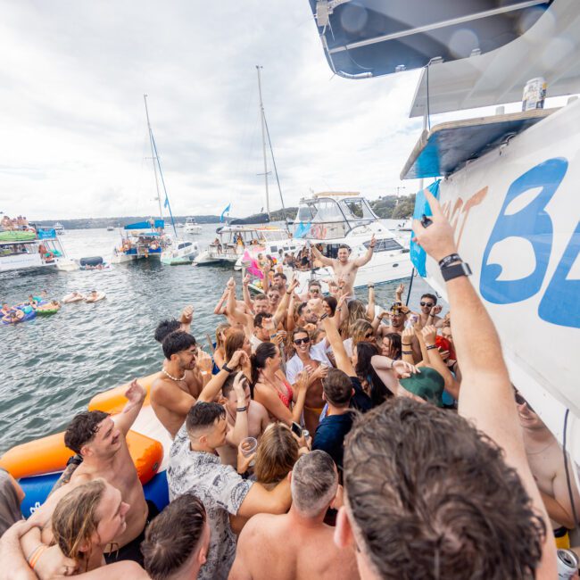 A large group of people partying on a yacht and in the water, with arms raised and drinks in hand. Several boats are anchored nearby. Some people are floating on inflatable rafts. The atmosphere is energetic and festive under a cloudy sky.