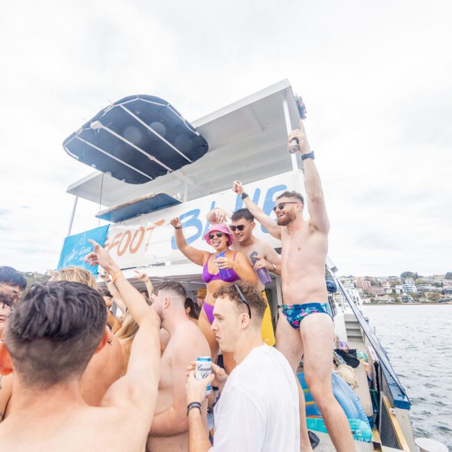 A group of people in swimsuits are on a sailboat, raising drinks and cheering. They are enjoying a sunny day on the water. The background shows a bright sky and distant buildings. The mood is festive and lively, perfect for making unforgettable memories.