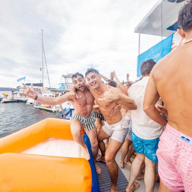 A group of people in swimwear enjoy a lively party on a yacht. Two men in the foreground pose playfully with wide smiles, making peace signs. Amidst other nearby boats, the scene is bursting with energy and fun. The cloudy sky adds a dramatic backdrop to this vibrant gathering.