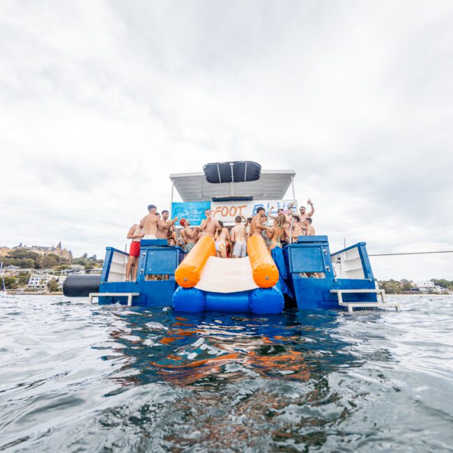 A group of people enjoys a day on the water with a large, blue floating structure and orange inflatable slides. They are gathered on the deck, some seated at the top of the slides, ready to descend into the water. Boats and land are visible in the background, while others relish an exciting floating party.