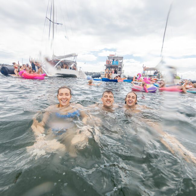 Three smiling people swim in the ocean surrounded by many others in inflatable tubes and boats during a lively carnival. The background shows several boats and a cloudy sky, with various activities happening on the water. The atmosphere is festive and energetic.