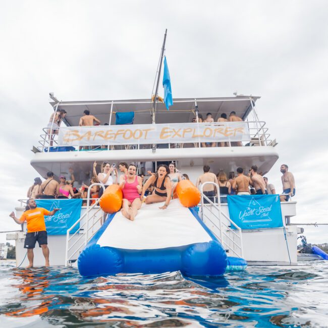 A group of people on a boat party. Two individuals are sliding down an inflatable water slide from the back of the boat into the water. The banner reads "BAREFOOT EXPLORER" and the boat is filled with people socializing and enjoying sunlit festivities.