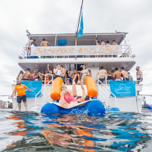 A group of people enjoy a lively boat party aboard a two-deck vessel named "Barefoot Explorer." The boat is adorned with flags and branded banners, adding to the festive atmosphere. One person is lounging on a large inflatable float in the water near the boat, creating a scene of perfect relaxation.