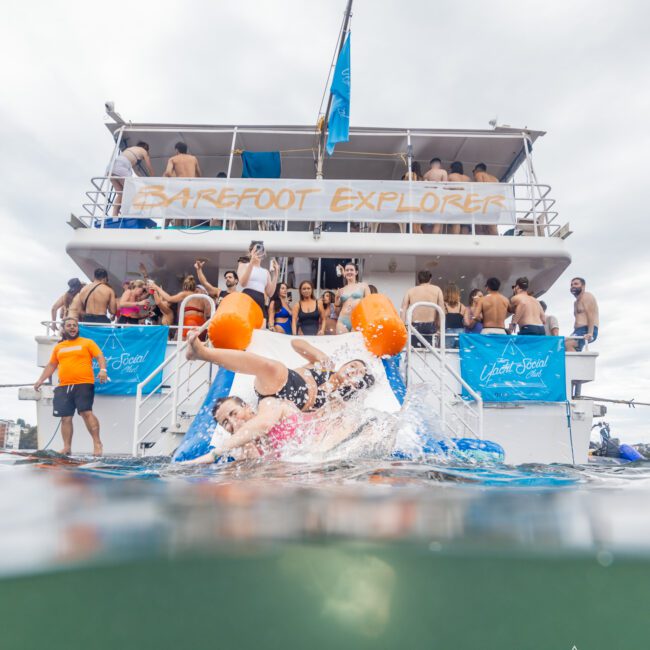 A large group of people is having fun on a two-story boat named "Barefoot Explorer" during one of the famous Boat Parties Sydney The Yacht Social Club. Some are diving into the water, while others are on the deck. The atmosphere is lively, with banners and life buoys visible. The logo of "The Yacht Social Club" is on a banner.