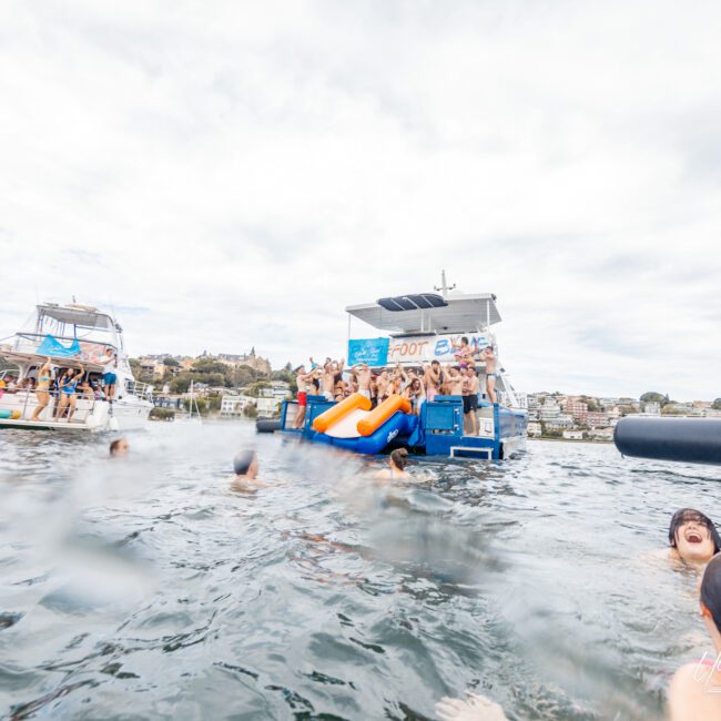 A group of people enjoy a sunny day on boats anchored near each other on a body of water. Some are on the boats, while others swim or float nearby. The scene is lively and festive, with music playing and an inflatable orange slide on one of the boats.