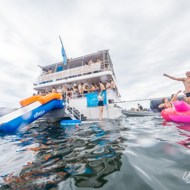 A lively group of people on a yacht named "Boat Foot Explorer" are enjoying a party, with some on the yacht and others on vibrant inflatable floats in the water. The atmosphere is festive under overcast skies. Text in the corner reads “The Yacht Social Club,” showcasing their adventurous spirits.