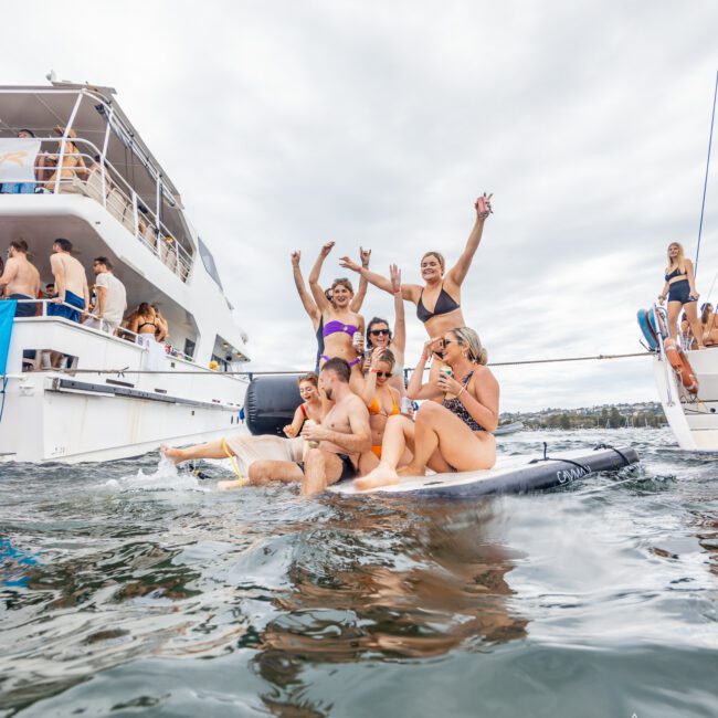 A group of people in swimsuits are having fun on a floating platform in the water near a large yacht. Some are sitting, while others are standing and waving their arms. The yacht has people on it enjoying the Yacht Social Club, and the sky is cloudy.