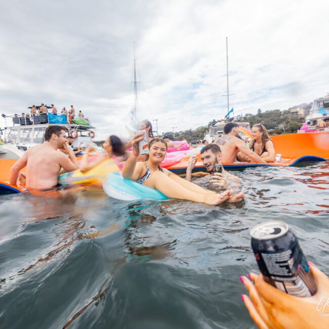 People are floating on colorful inflatables in the water near a couple of boats, enjoying the sunny weather. Some individuals are raising cans and smiling. The background shows a cloudy sky and additional people on the boats, creating a lively atmosphere.