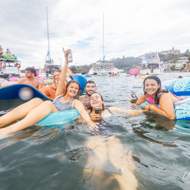 A group of people enjoying a sunny day in the water, floating on inflatable loungers, with boats and more people in the background. The group is smiling, holding drinks, and appears to be having fun. The scene captures a lively and social atmosphere, perfect for summer enjoyment.