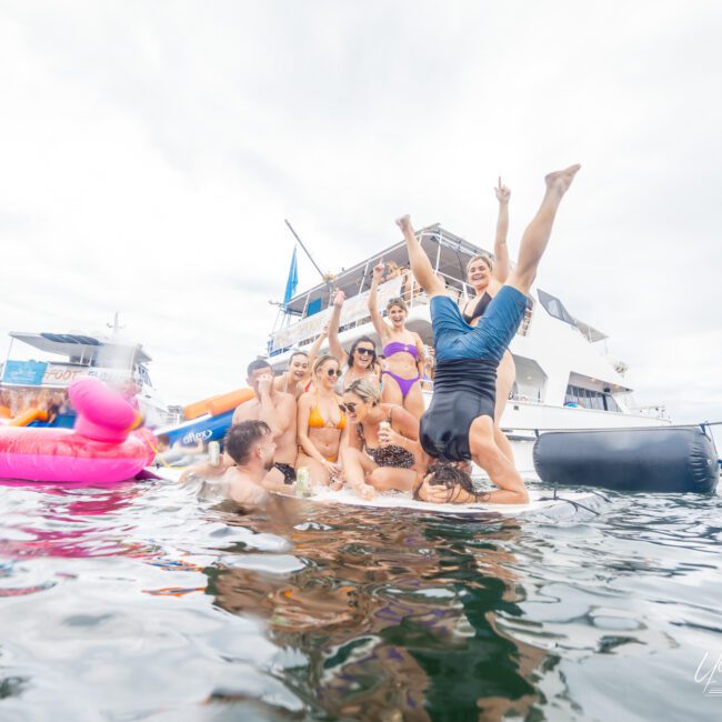 A cheerful group on a floating dock enjoys the day by the water near a boat. One person performs a handstand as others watch and smile. Inflatable pool toys, including a pink flamingo, float nearby, while the cloudy sky adds charm to the lively scene. The boat in the background has people on board enjoying themselves.