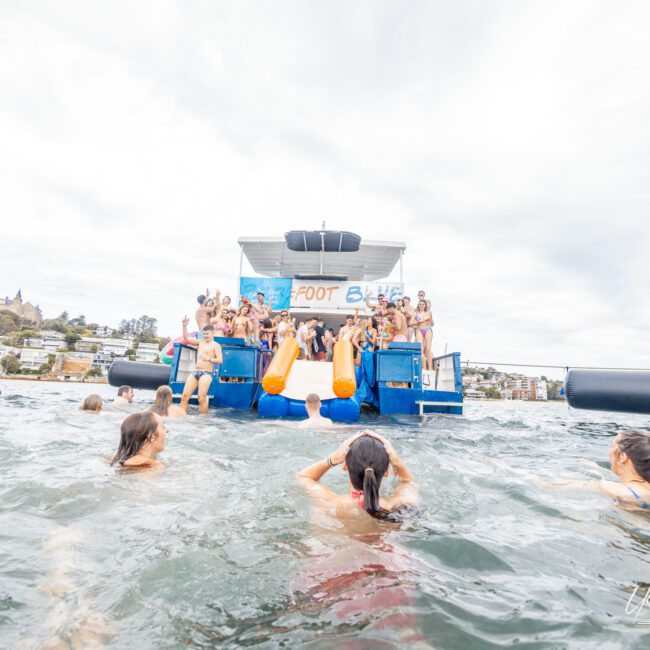 A group of people is celebrating on a floating platform with a banner that reads "AFLOAT." They are gathered and cheering, while several others swim nearby. The platform is anchored near the shore, with picturesque buildings and lush trees visible in the background, creating an idyllic scene.