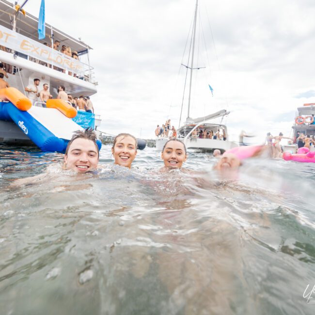 Four people smiling and enjoying the water near a boat named "Barefoot Explorer." There is a slide off the boat and people onboard, soaking up the sun. Another boat with more people is seen in the background on this perfect, sunny day.