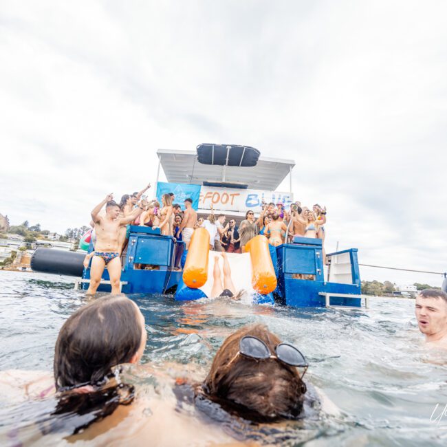 People enjoying a party on a large blue boat equipped with slides, floating on water. Several individuals are cheering and posing on the boat, while others are swimming nearby. The lively atmosphere is enhanced by cloudy skies in the background and the vibrant music playing from onboard speakers.