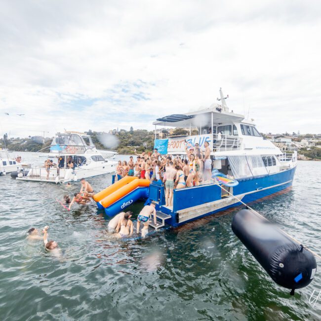 A large group of people are enjoying a party on and around a blue and white boat anchored in the water. Some are swimming or floating on inflatables, while others are dancing on the boat's deck. Other boats and a picturesque coastline with houses are visible in the background.