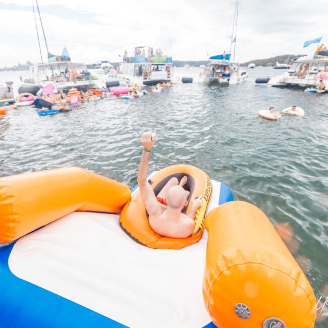 A person lounges on a large orange and white inflatable in the water, surrounded by numerous boats and people swimming or floating on inflatables. It appears to be a lively, social scene with bubble enthusiasts enjoying the sunny day.