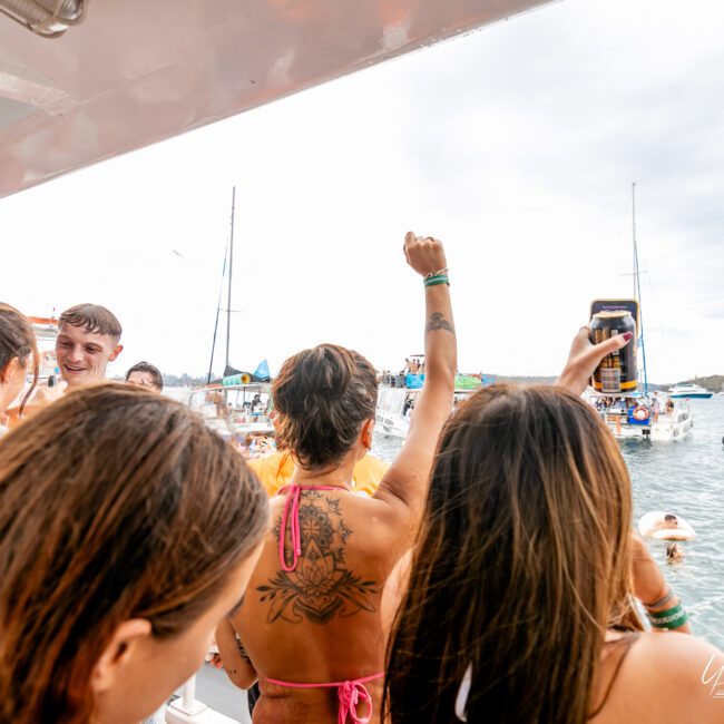 A group of friends on a boat party, some holding drinks, with one person raising their arm in celebration. The boat is anchored near other boats on the water, with people swimming and enjoying the serene weather.