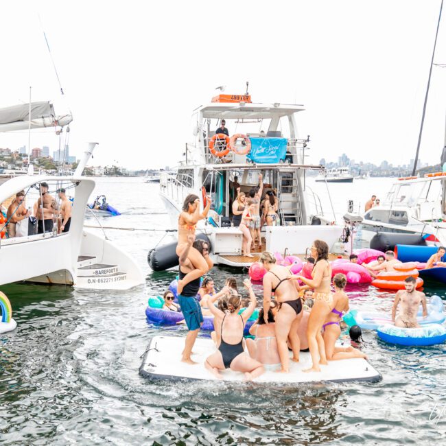 A lively outdoor party is taking place on the water with people on various inflatables and a floating platform. Boats surround the area, and attendees are enjoying music, dancing, and socializing in swimsuits under a partly cloudy sky. The festive atmosphere radiates joy and fun.