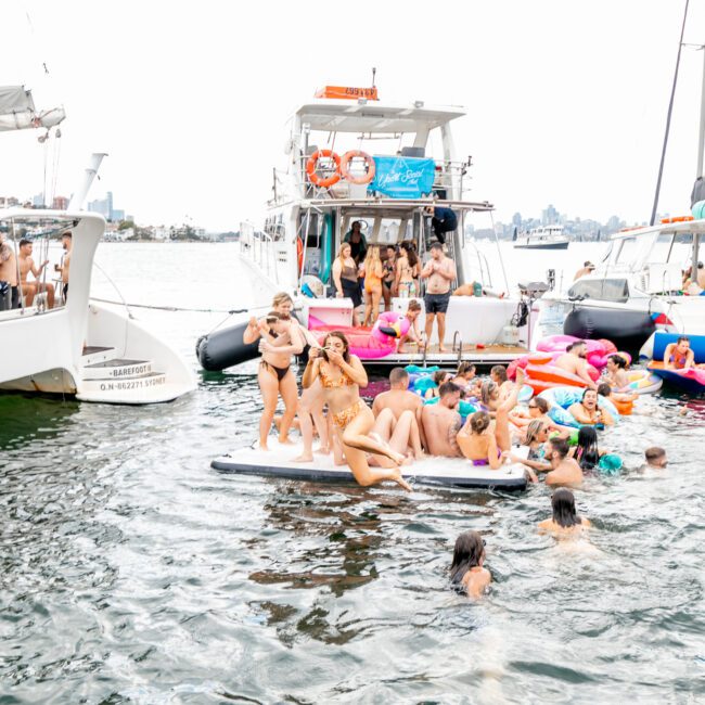 A lively scene of people enjoying a boat party on the water. Many are on large inflatable floats, swimming, and mingling around the boats, while others gather on the decks. The background features distant city buildings under a bright sky with vibrant water reflections adding to the festive atmosphere.