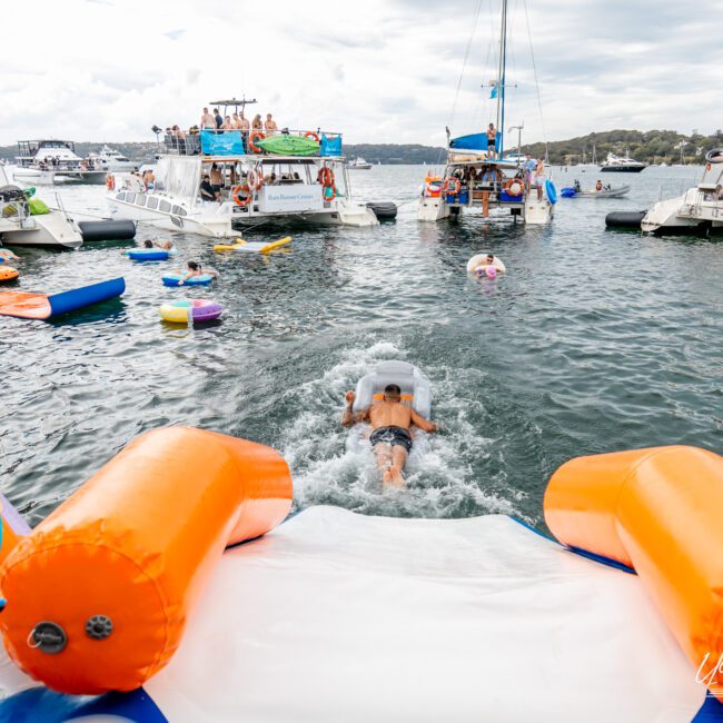 A person slides down an inflatable water slide into a calm body of water. Several boats and yachts are anchored nearby with people enjoying the day both onboard and in the water. Inflatable floaties and colorful pool toys dot the surface, creating a festive atmosphere under the slightly cloudy sky.