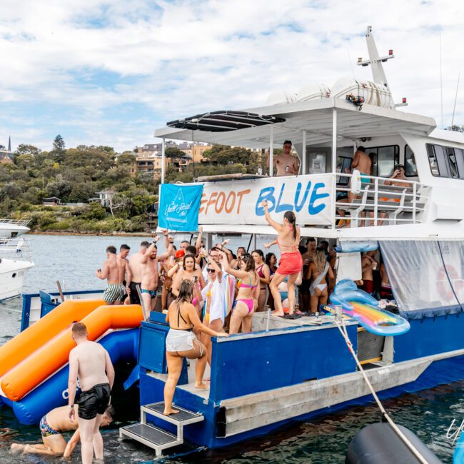 A group of people in swimsuits are having a lively party on a boat named "Foot Blue" equipped with a water slide and vibrant music. The boat is anchored in a scenic area with other boats and houses visible in the background. Everyone appears to be enjoying the sunny day.