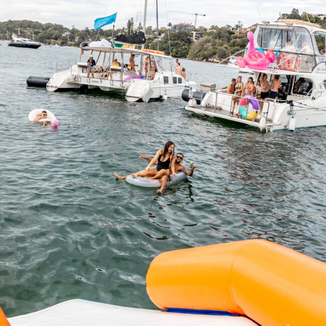 A fun scene on a lake with people partying on anchored boats. In the foreground, a couple is lounging on a float in the water. Inflatable toys and slides are also visible, with more people enjoying the festivities. The background includes an idyllic shoreline framed by lush greenery and crystal-clear waters.