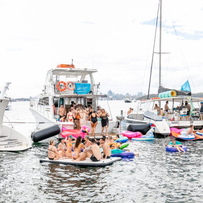 A group of people are gathered on boats and inflatables in a body of water. Several boats are anchored close together, with people mingling and relaxing on the decks and floating on colorful inflatables. The overcast sky adds a calm atmosphere, and buildings are visible in the distance.