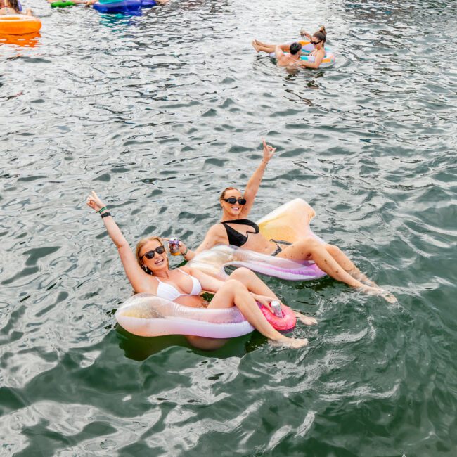 Two women in bikinis relax on inflatable pool floats in a body of water, raising their arms in a celebratory gesture. Behind them, people on other floats and yachts enjoy the sunny day. Logo at the bottom right reads "Yacht Social Club.