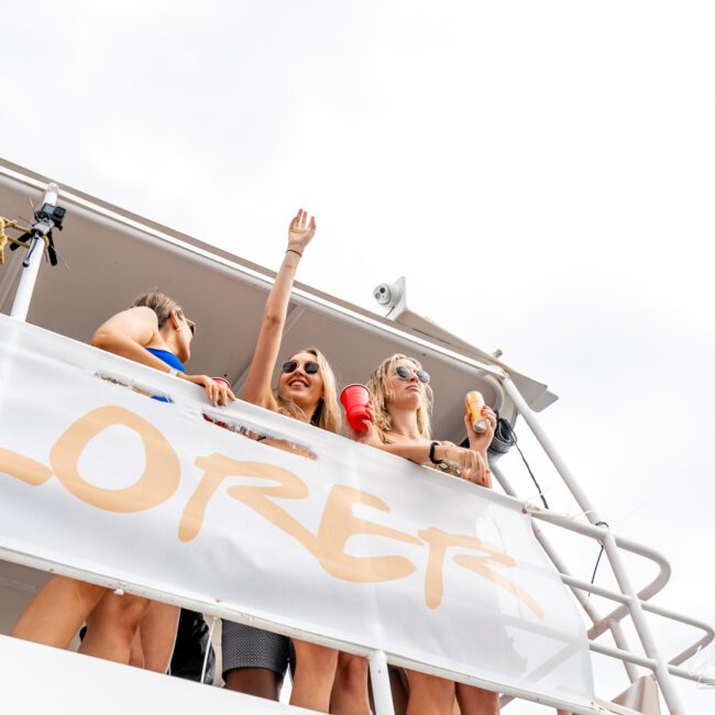 Four people smiling and enjoying themselves on the deck of a boat, with two women holding beverages and waving. A GoPro is attached to the railing, capturing the fun moments, and a large banner saying "EXPLORER" is visible. The sky is clear in the background.