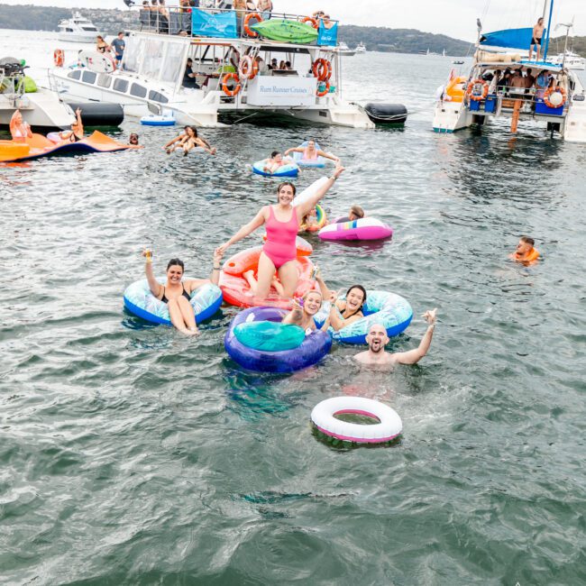 A lively group of people enjoy a day basking in the sun on colorful inflatable rings, smiling and cheering. Nearby, boats are anchored close together as more people socialize under a clear blue sky. The festive scene is bustling with energy and fun.