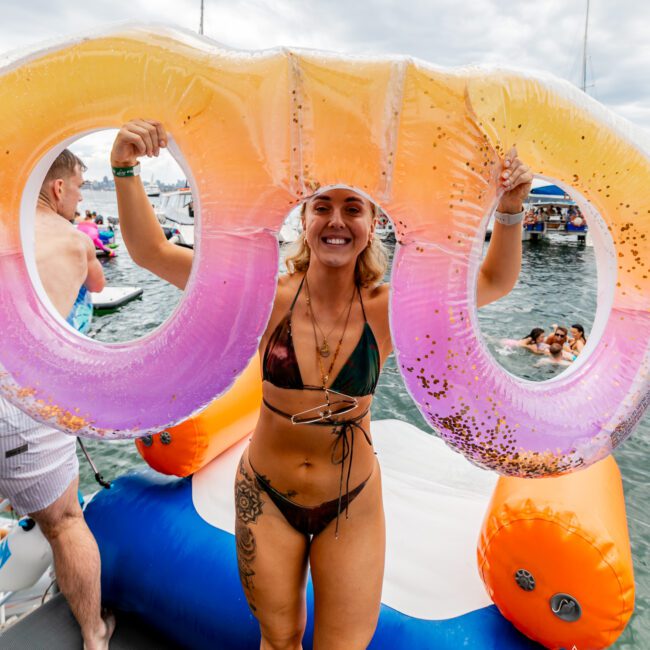 A woman in a bikini stands on a dock near water, holding a large, colorful floatation device shaped like eyeglasses above her head. She is smiling and there are people swimming and boats in the background. The scene appears to be part of a lively summer gathering.