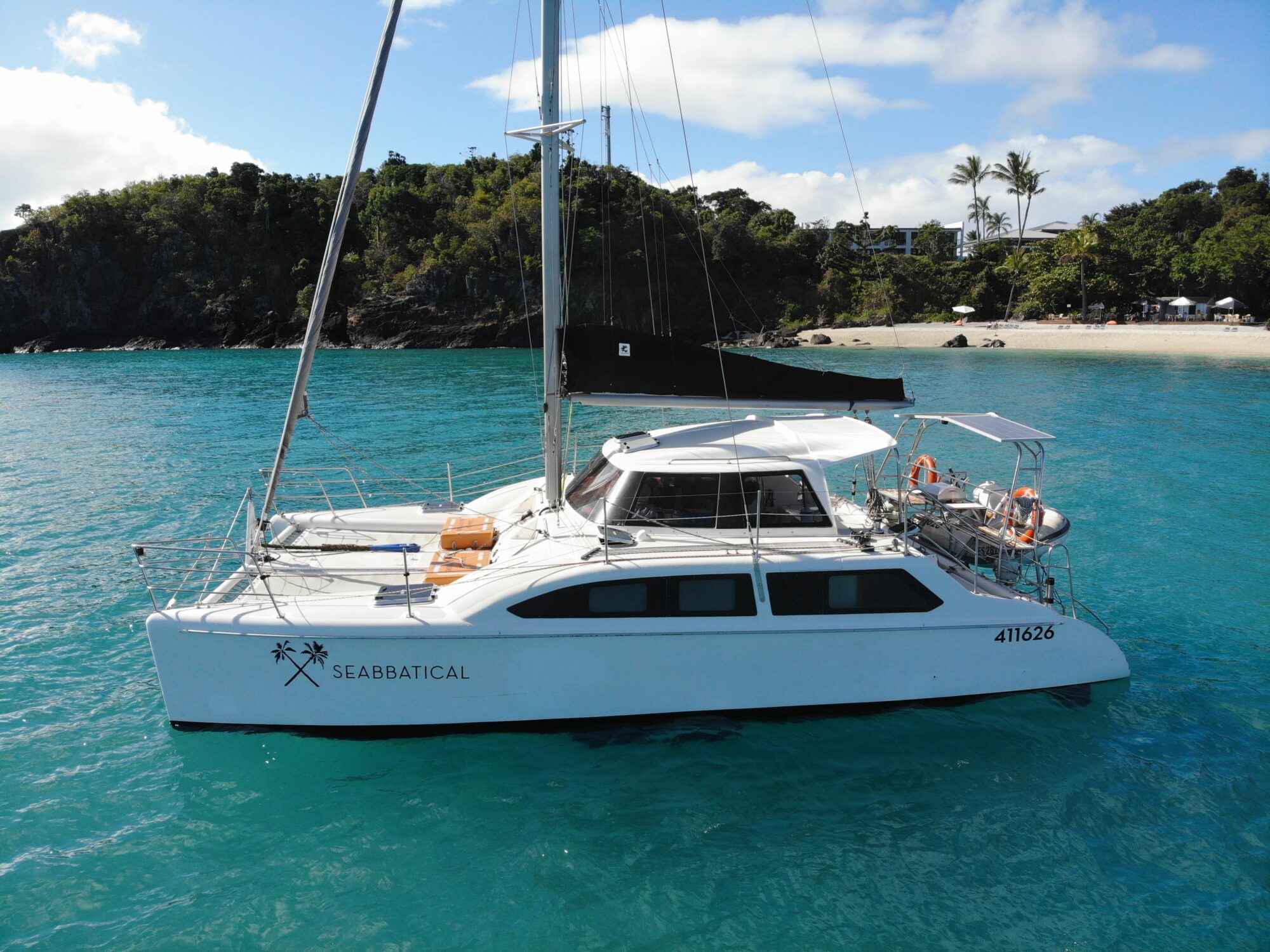 A white catamaran sailboat named "Seabbatical" is anchored in clear blue waters near a lush, green shoreline with a sandy beach. The boat, perfect for those seeking an adventure, has outdoor seating and is equipped with lifebuoys. Palm trees and a few structures are visible in the background.