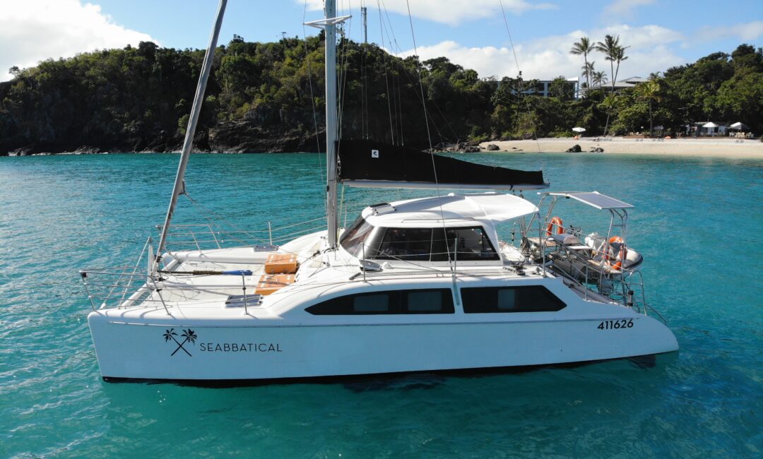 A white catamaran named "Seabbatical" is anchored in clear turquoise water near a lush, green island with a sandy beach. The vessel's registration number is 411626. The sky is partly cloudy, and the scene is serene and tropical, perfect for an island getaway.