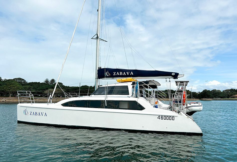 A white sailboat named "Zabava" floats on calm waters. The boat boasts a black sail cover displaying the name 'Zabava'. Tall trees and a partly cloudy sky create a serene backdrop, while a small dinghy is securely fastened at the back of the sailboat, adding to its charm.