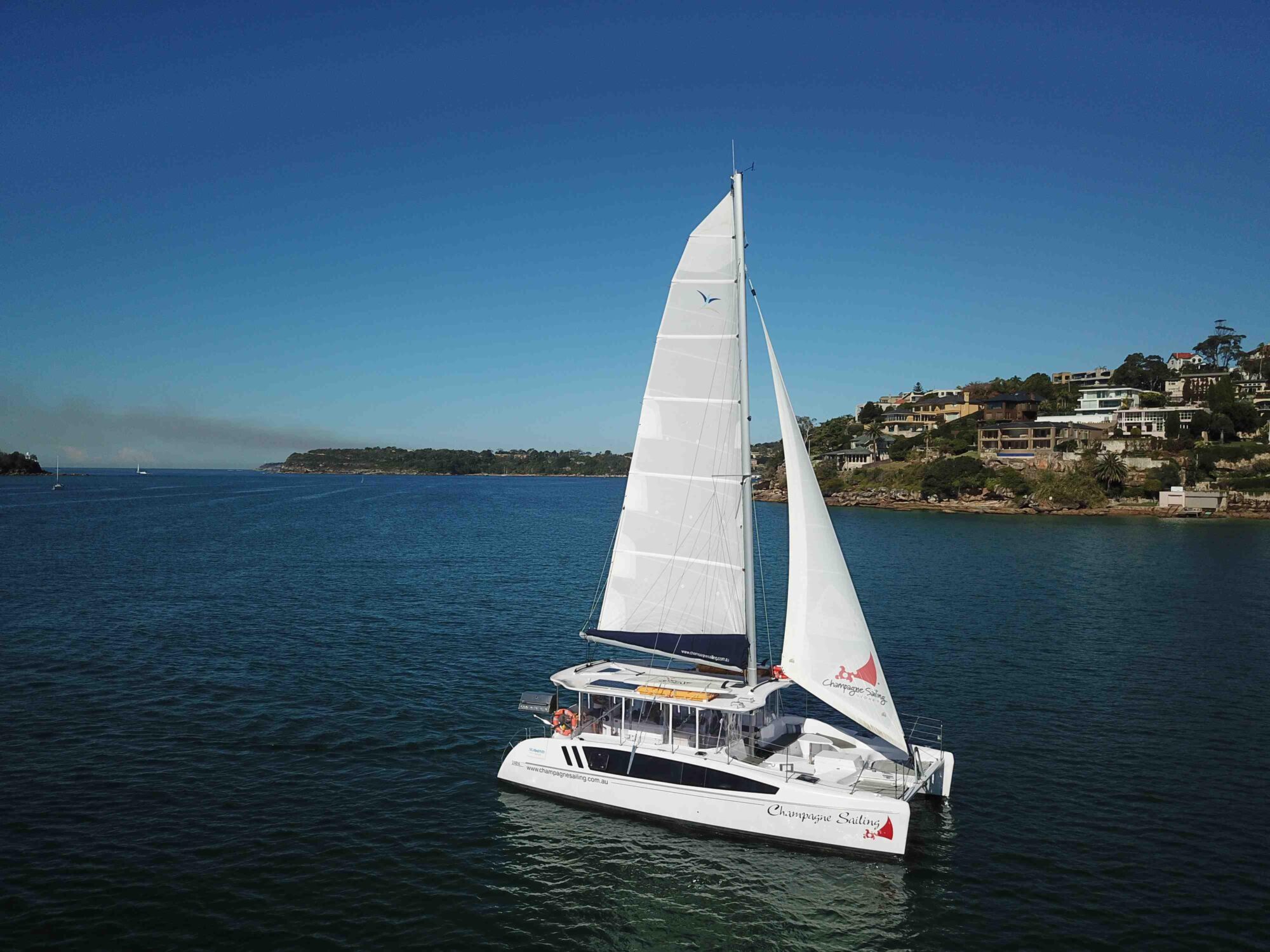 A white catamaran named "Escape Series" with its sails up navigates calm, azure waters near a coastal area boasting lush, green hills and several charming houses. The sky is clear and blue, indicating beautiful, sunny weather.